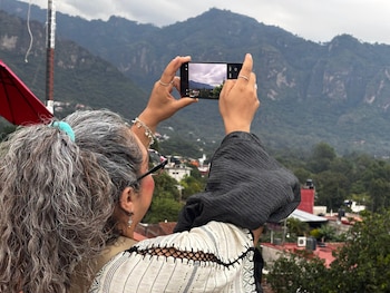 Vista trasera de una mujer con cabello gris rizado sujetando un teléfono móvil para fotografiar un paisaje montañoso con casas