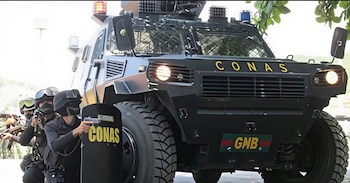 Un vehículo blindado oscuro con las siglas CONAS en amarillo y una placa GNB, junto a oficiales con equipo táctico y armas