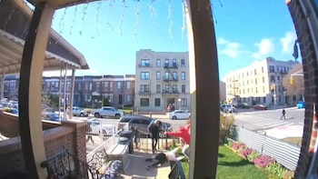 Desde un porche, se ve a una persona caída y otra de pie sobre ella. Al fondo, calle con coches y edificios de varios pisos bajo un cielo azul