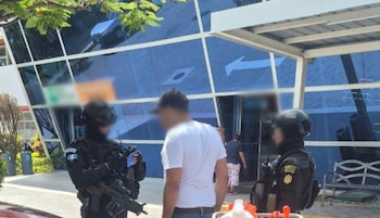 Dos agentes uniformados con cascos e instrumental táctico conversan con un hombre vestido con camiseta blanca junto a un edificio de vidrios azules
