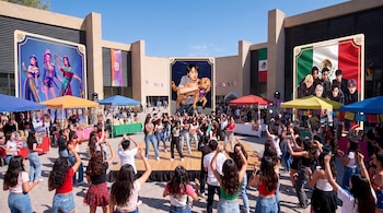 Jóvenes bailan sobre una tarima de madera en una plaza rodeada de carpas de colores y carteles decorativos gigantes.