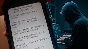 Primer plano de un teléfono mostrando una alerta de emergencia de tsunami en Perú; al fondo, un hacker encapuchado teclea en una laptop con código en pantalla.