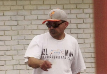 Un hombre con gorra deportiva, anteojos de sol y camiseta blanca estampada posa frente a una pared de ladrillos pintados de blanco