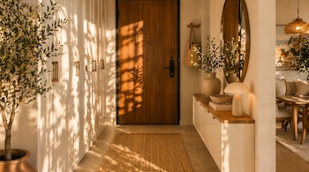 Entrada de casa con una puerta de madera oscura, paredes blancas, planta grande en maceta de terracota, y un banco con espejo y lámpara. La luz del sol proyecta sombras.