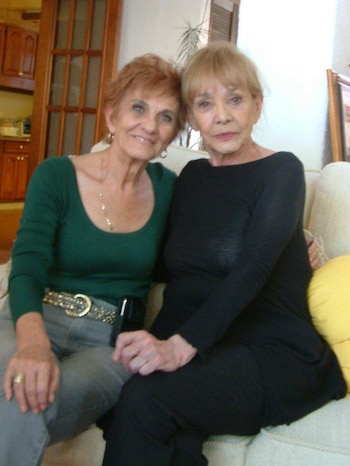 Dos mujeres, una con cabello rojizo y camiseta verde, sonríe junto a otra de cabello rubio y blusa negra, sentadas en un sofá claro