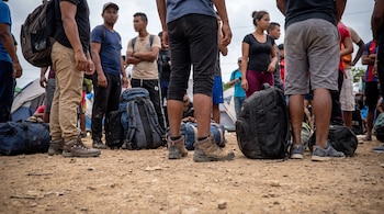 Un grupo de personas de pie sobre tierra, con mochilas y bolsas de lona en el suelo; tiendas de campaña se observan en el fondo.