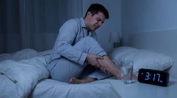 Hombre en pijama con expresión de dolor en la cama, sujetando su pantorrilla. En la mesita de noche hay un vaso de agua y un reloj digital marcando 3:17 AM.