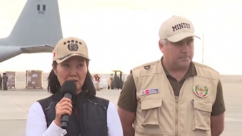 Mujer con micrófono y gorra con escudo de Perú, hombre con gorra MINDEF y chaleco Ministerio de Defensa. Fondo: avión militar, cajas y personal en pista de aeropuerto