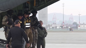 Un hombre detenido desciende de la rampa de una aeronave escoltado por agentes policiales y militares armados.