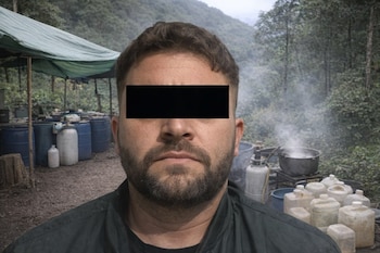 Primer plano de un hombre con el rostro censurado, de pie frente a un supuesto narcolaboratorio en la sierra con barriles azules, ollas humeantes y recipientes plásticos.