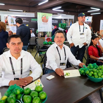 Dos hombres sentados en una mesa con limas frescas en bolsas de malla y un cuenco, sosteniendo folletos en un evento agrícola con banners de fondo