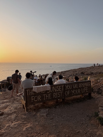 Varias personas en un acantilado rocoso. Un banco de madera con texto, mesas y sillas. El sol poniente en el horizonte sobre el mar