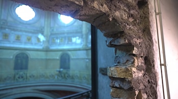 Pared de piedra y ladrillo expuesto que enmarca una vista al interior de un edificio con cúpula y ventanas circulares.