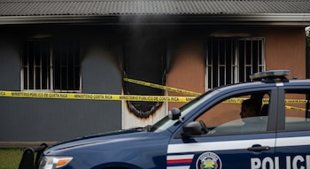 Vivienda con fachada quemada y humo visible, acordonada por cinta amarilla del Ministerio Público de Costa Rica. Patrulla policial azul en primer plano.