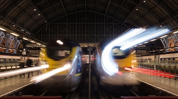Estación de trenes nocturna con cubierta metálica, andenes y dos trenes frontales que dejan estelas luminosas por exposición prolongada.