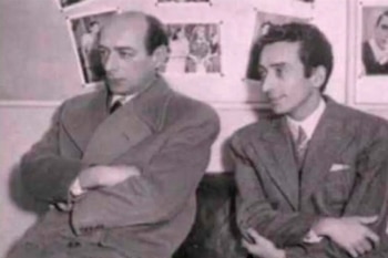 Dos hombres vestidos con traje están sentados con los brazos cruzados frente a un panel con varias fotografías en blanco y negro
