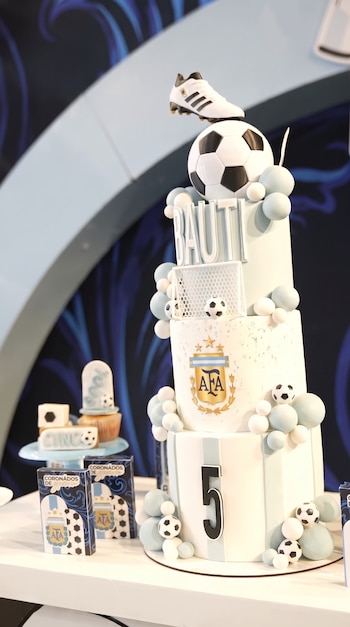 Un pastel de varios pisos decorado con una bota de fútbol, un balón, una portería, el nombre Bauti, el número cinco y el logo de AFA, junto a cupcakes y cajas.