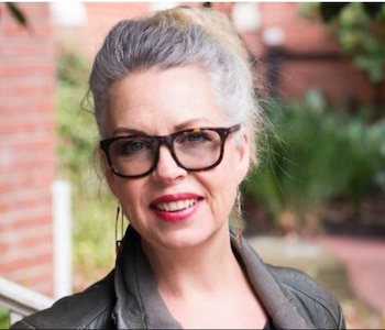 Primer plano de una mujer con cabello gris recogido, gafas de montura oscura y labios rojos. Sonríe a la cámara. Viste una chaqueta oscura. Fondo exterior borroso