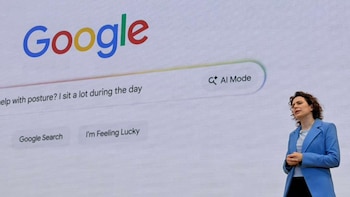 Una mujer de cabello rizado y blazer azul claro habla frente a una pantalla blanca con el logo de Google, una barra de búsqueda y el botón 'AI Mode'