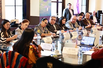 Varias personas, hombres y mujeres, sentadas alrededor de una mesa de cristal en una sala de conferencias con botellas de agua, documentos y una laptop
