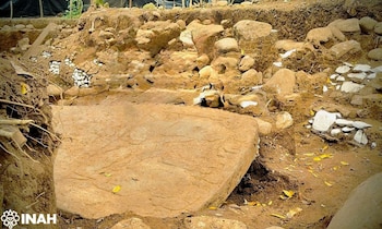 Una excavación muestra una losa de piedra con grabados sobre tierra, rodeada de rocas, escombros y el logo de INAH en la esquina inferior izquierda