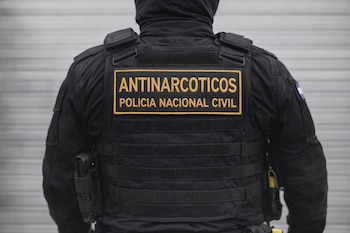Vista trasera de un oficial de policía vestido de negro, con un chaleco táctico que dice "ANTINARCOTICOS POLICIA NACIONAL CIVIL" en letras doradas.