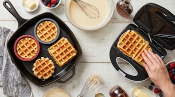 Waffles en una waflera y en una sartén con moldes. Mesa de madera clara con masa, bayas, mantequilla y jarabe, y una mano sacando un waffle.