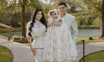 Fotografía de Karely Ruiz, un hombre y un bebé, todos vestidos de blanco, en un sendero exterior con árboles y un estanque al fondo