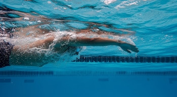 Nadador visto bajo el agua extendiendo un brazo hacia adelante al desplazarse por una piscina, con burbujas y líneas de carril visibles.