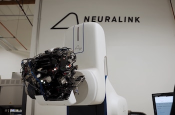 Máquina robótica blanca con brazos articulados y componentes internos negros y metálicos expuestos, frente a una pared con el logo de Neuralink