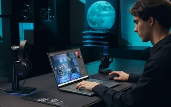 Un joven sentado frente a una computadora portátil gaming abierta en un escritorio oscuro, con auriculares en un soporte y un micrófono cerca