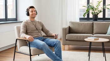 Un hombre de unos 30 años con auriculares de cable negros está sentado sonriendo en un sillón beige en un living limpio y ordenado, sosteniendo un móvil.