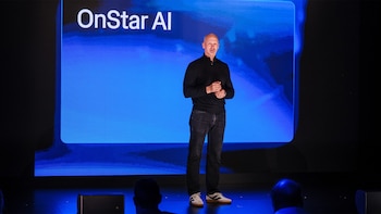 Un hombre de pie sobre un escenario frente a una pantalla azul con el texto OnStar AI, con espectadores en primer plano