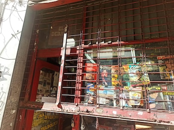 Vista frontal de un quiosco de periódicos y revistas cerrado con una reja metálica roja oxidada. Se ven revistas antiguas de deportes, cultura y cómics tras el cristal