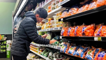 Un hombre vestido con una chaqueta negra con capucha mira productos preenvasados, incluyendo ensaladas y zanahorias, en el estante refrigerado de un supermercado