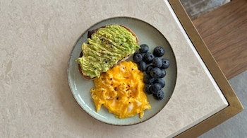 Vista aérea de un plato con una tostada de aguacate, huevos revueltos espolvoreados con pimentón y un montículo de arándanos frescos sobre una mesa de mármol.