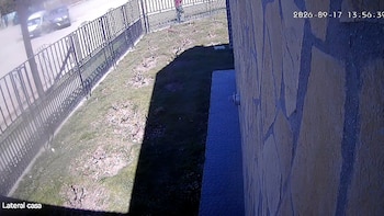 Toma de cámara de seguridad que muestra una reja metálica negra, un patio con césped, una pared de piedra y automóviles en una calle