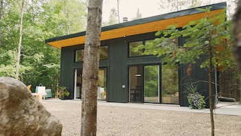 Fachada de una casa de madera oscura con ventanales de vidrio, alero de madera clara y suelo de gravilla entre árboles