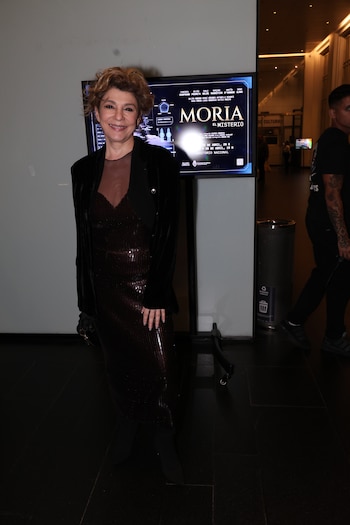 Carolina Papaleo sonríe, vestida con un vestido largo de lentejuelas oscuras y chaqueta de terciopelo, frente a un cartel digital de "Moria El Misterio"