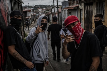 Un grupo de jóvenes con el rostro cubierto con pañuelos y gorras posan en una calle angosta entre casas.
