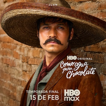 El actor Louis David Horné, con sombrero de ala ancha, bigote y barba, mira directamente al frente. Viste una chaqueta de cuero marrón y un pañuelo rojo, posando contra una pared de ladrillo