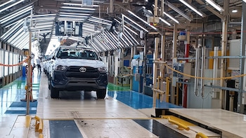 Una camioneta pickup Toyota blanca avanza por una línea de montaje industrial bajo intensas luces fluorescentes, con equipo de fábrica y un trabajador en el fondo