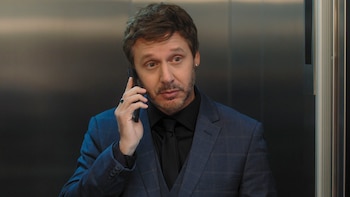 Hombre de mediana edad con barba y cabello castaño, vestido con un elegante traje a cuadros azul marino y camisa negra, hablando por un teléfono celular negro