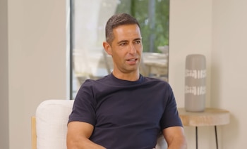 Hombre sentado en una silla, viste camiseta azul oscuro, en una sala con ventana, mesa auxiliar y objeto decorativo gris