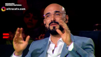 Un hombre calvo con barba, anteojos y saco gesticula con las manos levantadas ante un fondo oscuro