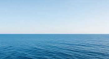 Fotografía de una vasta extensión de océano azul con pequeñas olas, que se encuentra con un cielo azul pálido y despejado en el horizonte recto y nítido.