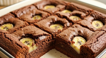 Primer plano de un brownie de banana y cacao en una fuente cuadrada, cortado en nueve porciones, con rodajas circulares de plátano visibles en la superficie.