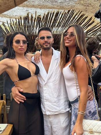 Tres personas, un hombre y dos mujeres, posan al aire libre bajo una estructura de madera. El hombre lleva camisa blanca y gafas de sol, las mujeres llevan atuendos ligeros y gafas de sol