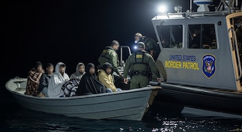 Operativo nocturno de la Patrulla Fronteriza de EE. UU. Una yola con migrantes sentados y cubiertos con mantas es interceptada por una lancha oficial en aguas oscuras.