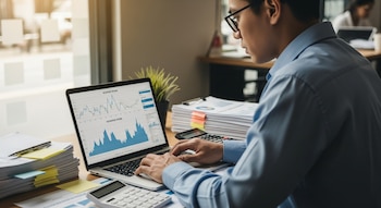 Un hombre usa una laptop con gráficos de datos, junto a pilas de documentos, notas, calculadoras y una planta en un escritorio de oficina.